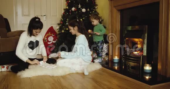 快乐的两个姐姐装饰圣诞节和新年树和可爱的儿子吃苹果和看相机靠近壁炉视频的预览图