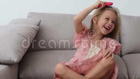 六岁的快乐女孩穿着粉红色的裙子坐在沙发上梳着金发视频的预览图