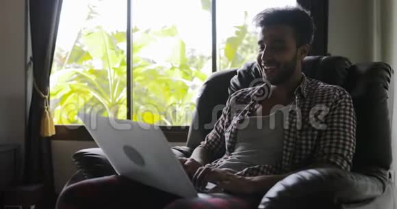 男人坐在扶手椅上用笔记本电脑打字在家工作男人上网快乐微笑视频的预览图