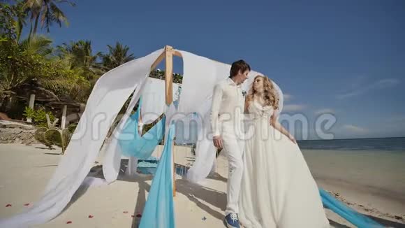 在海边的热带海滩上结婚在拱门下亲吻白色和蓝色的翅膀在空中飞翔视频的预览图