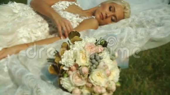 美丽的金发新娘穿着婚纱躺在靠近婚礼花束的草地上视频的预览图