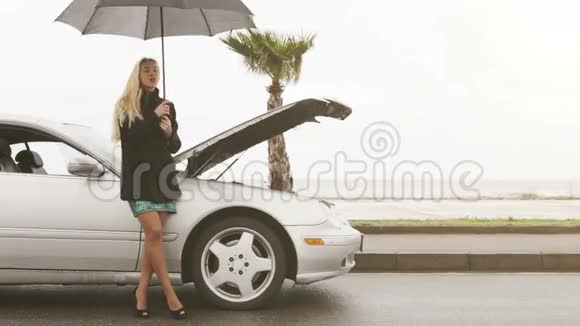 一个年轻的金发女孩站在伞下的破车旁视频的预览图