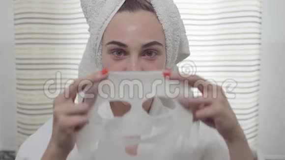 一个年轻的开朗的女人在镜头里在浴室里把床单面具戴在脸上可爱的女孩与不同视频的预览图