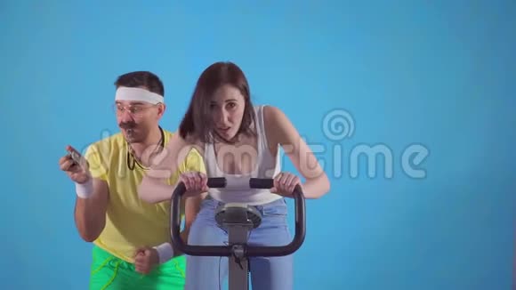 有趣的男子教练80带胡子训练一个年轻的女人骑着蓝色背景的慢摩运动自行车视频的预览图