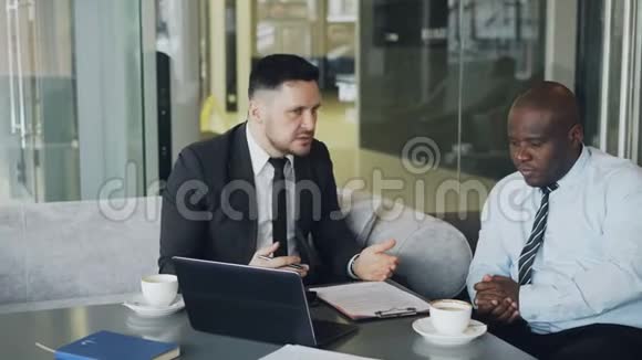 穿黑色西装的愤怒商人在现代咖啡馆开会时严厉批评了他的非裔美国雇员视频的预览图