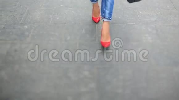 穿着牛仔裤和时髦红鞋的时尚年轻女士穿过人群走在城市街道上视频的预览图