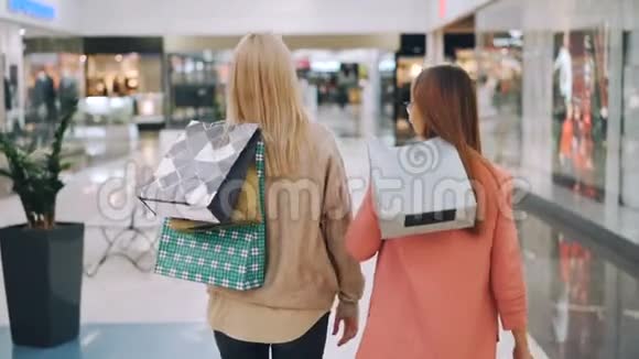 快乐的年轻女性朋友们在购物中心手里拿着明亮的袋子走在一起然后转向相机视频的预览图
