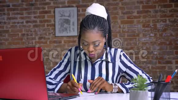集中精力的非洲妇女写笔记坐在工作场所附近的红色笔记本电脑砖背景视频的预览图