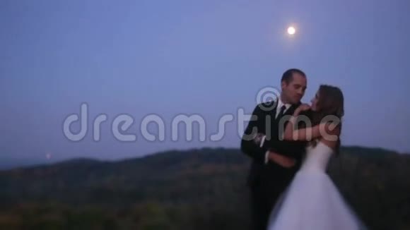 美丽的年轻夫妇迷人的新娘靠在可爱的新郎的肩膀上晚上在公园户外接吻视频的预览图