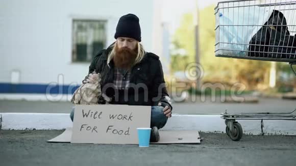年轻人帮助无家可归的人给他一些钱而乞丐喝酒坐在购物车附近视频的预览图