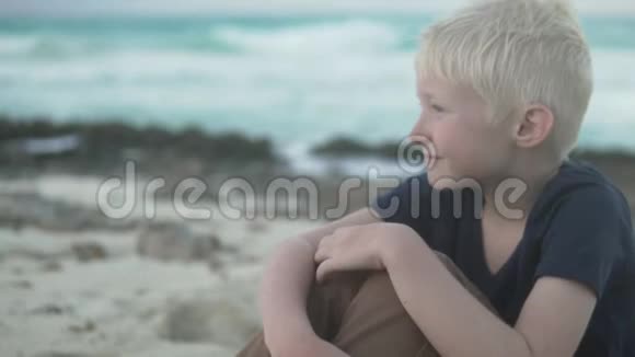 一个金发男孩坐在海边的沙滩上视频的预览图