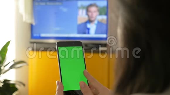一个女人正在看电视拿着一台绿色屏幕的智能手机在电视节目上的新闻视频的预览图