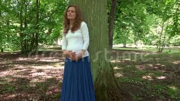 快乐的白种人怀孕的女孩抚摸着她的腹部站在大树干附近视频的预览图