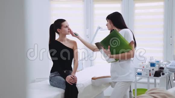 年轻的黑发女人穿着优雅的衣服坐在美容师的柜子里美容师检查她的脸他们视频的预览图