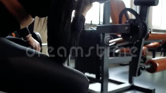 穿着黑色紧身裤和上衣的年轻女孩为二头肌做运动坐在长凳上在健身房举起哑铃视频的预览图