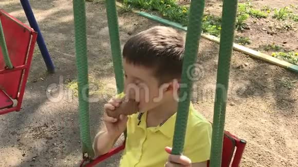 一个十几岁的白种人男孩坐在秋千上在孩子们夏天的操场上吃着美味冰淇淋视频的预览图