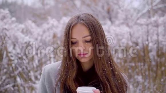 在冬天的公园里年轻美丽的悲伤女人端着一次性咖啡或茶杯坐在外面的长凳上视频的预览图