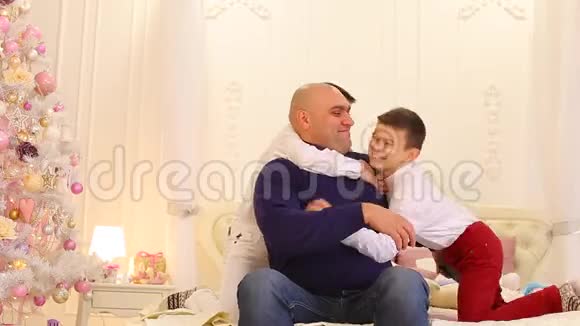 快乐的父亲和双胞胎兄弟的孩子在明亮的卧室里的大床上有圣诞树视频的预览图