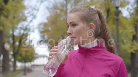 一位穿着粉红色运动服的年轻白种人女人喝着水逃跑的特写肖像自信的女选手视频的预览图