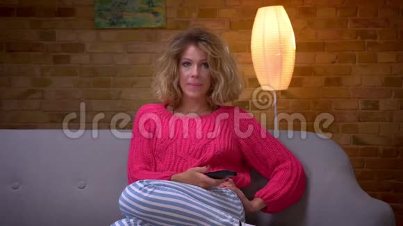 金发的家庭主妇穿着粉红色毛衣在沙发上看电视吓坏了把她的脸藏在舒适的家里视频的预览图