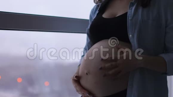 一个快乐的孕妇站在一个大的全景窗口位于一个高空他摸他的肚子视频的预览图
