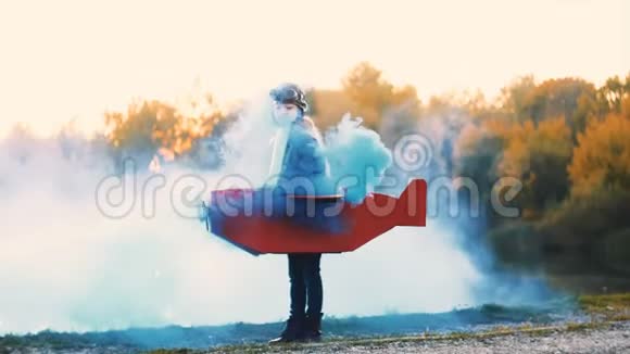 快乐的小飞行员女孩站在湖边穿着纸板飞机服装蓝色烟雾播放飞行员慢动作视频的预览图