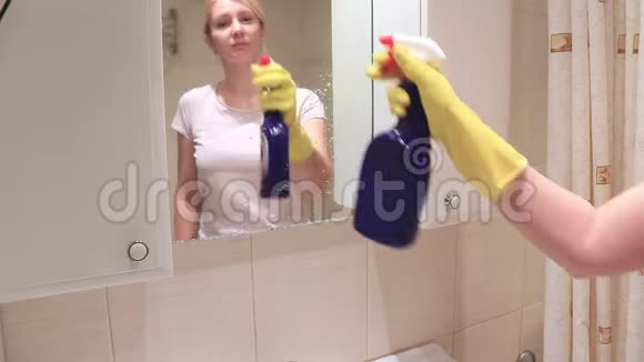 年轻的金发女人戴着黄色的橡胶手套洗着浴室的镜子洒着雨刷喷雾和泡沫清洁剂清洁雨刷视频的预览图
