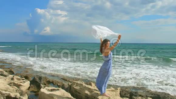 美丽的女士穿着长裙在岩石海滩上挥舞着白色的围巾视频的预览图