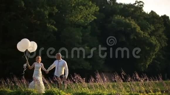 快乐的年轻新婚夫妇在夕阳下的夏日田野上带着气球散步浪漫婚礼概念视频的预览图