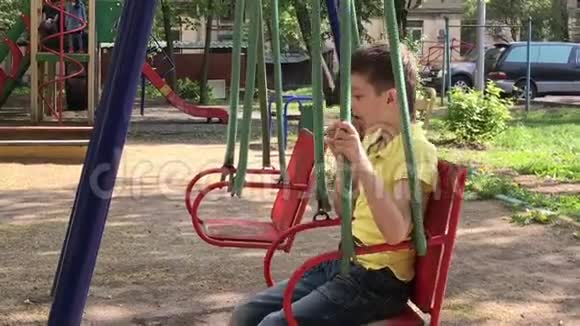 一个十几岁的白种人男孩坐在秋千上在孩子们夏天的操场上吃着美味冰淇淋视频的预览图