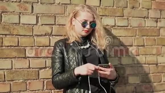 时尚的金发女郎戴着太阳镜和皮夹克用手机在蓝牙耳机上听音乐视频的预览图