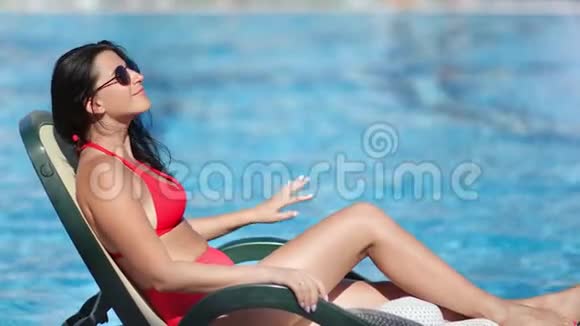 穿着红色比基尼的舒适黝黑的旅行女性在酒店游泳池附近的躺椅上放松视频的预览图