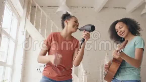 混合种族的年轻滑稽女孩跳舞用吹风机唱歌在床上弹声吉他姐妹们玩得开心视频的预览图