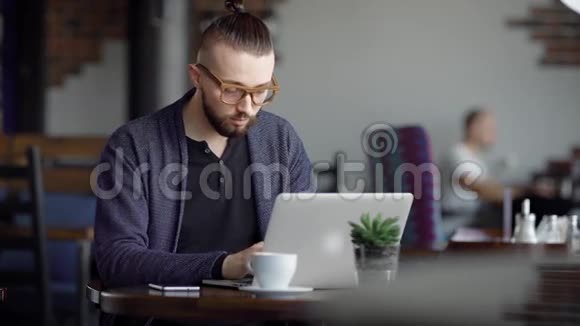 用现代笔记本电脑把一个戴着眼镜黑色t恤和蓝色夹克的英俊男子关在咖啡馆里男作家视频的预览图