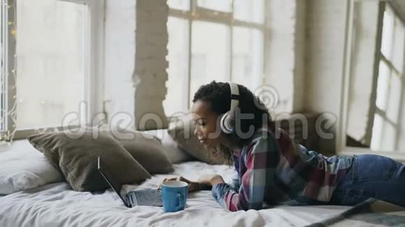迷人的混血女孩一边听音乐一边在床上笔记本电脑上浏览社交媒体视频的预览图