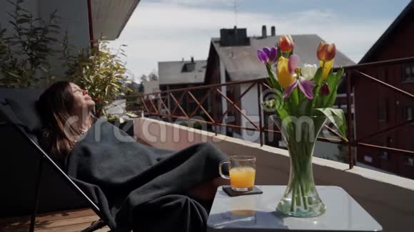 快乐的微笑着穿着格子布的年轻女子在阳台上享受着春天的阳光一束彩色郁金香和一杯橙汁视频的预览图