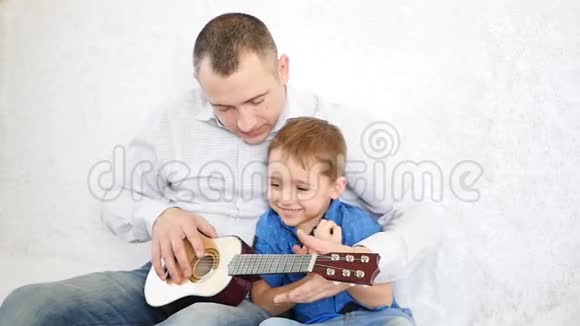 快乐的爸爸教儿子在白色沙发上弹吉他幸福家庭的概念视频的预览图