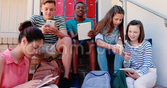 使用手机和数字平板电脑在楼梯上微笑的学校朋友小组视频的预览图
