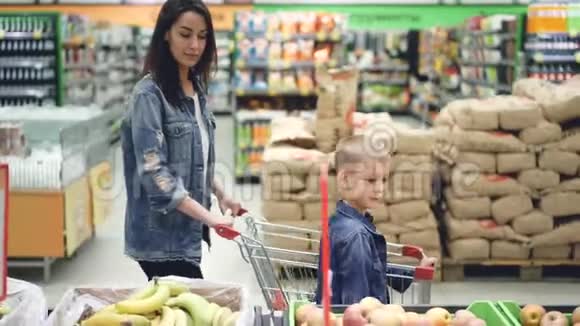 家庭欢乐的侧面景色妈妈和儿子在超市里散步用购物手推车选择水果和交谈视频的预览图