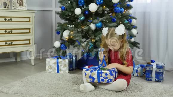 一个穿蓝色连衣裙的小女孩在圣诞树下打开新年礼物视频的预览图