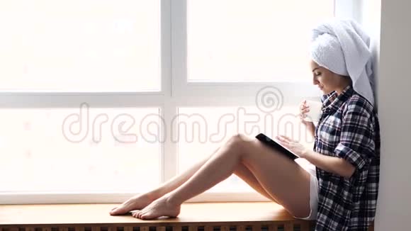 年轻女子在检疫期间呆在家里洗澡后坐在窗口看书喝茶或咖啡享受视频的预览图