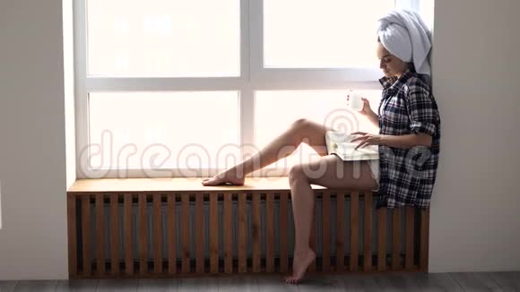 年轻女子在检疫期间呆在家里洗澡后坐在窗边看书独自喝茶或咖啡视频的预览图