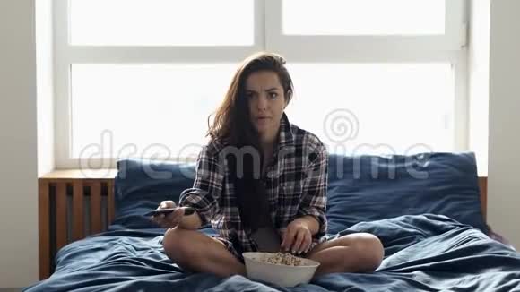 年轻女子在检疫期间呆在家里女孩坐在床上害怕看电影吃爆米花换电视视频的预览图