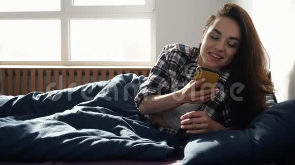 年轻女子在检疫期间呆在家里在智能手机屏幕上键入电话号码并愉快地交谈躺在床上视频的预览图