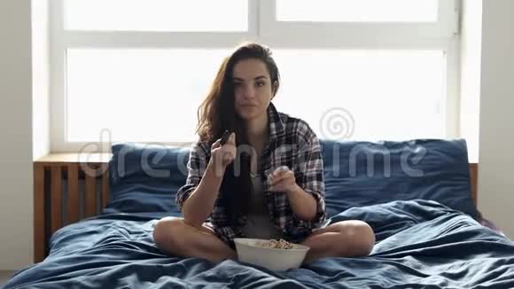 隔离期间年轻女子呆在家里女孩坐在床上换电视频道吃爆米花有点害怕视频的预览图