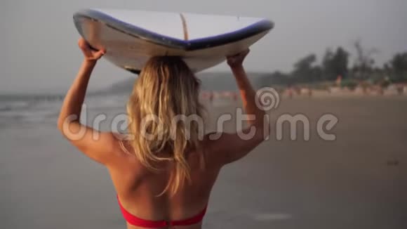 穿着红色泳衣的漂亮年轻女子带着冲浪板沿着海滩散步视频的预览图