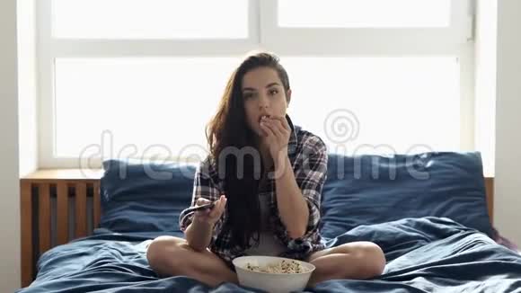 年轻女子在检疫期间呆在家里独自坐在床上看电视或电影吃爆米花使用遥控器视频的预览图