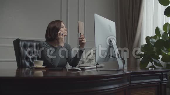 年轻的女商人在智能手机上聊天布鲁内特在办公桌前工作电脑显示器坐在扶手椅上视频的预览图
