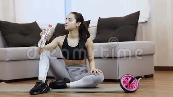 年轻女子在隔离期间在家锻炼独自坐在瑜伽垫上从瓶子里喝水休息并拥有视频的预览图