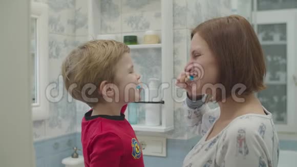年轻的母亲和穿着红色睡衣的学龄前男孩刷牙有趣的家庭沐浴常规妈妈和儿子一起笑视频的预览图